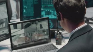 Tek kulaklı bir kulaklıkla konuşan erkek siber güvenlik memurunun omzunun üstünden. Bu arada bilgisayar ekranında birden fazla tek renkli CCTV kameraları izliyor ve gece ofisinde çalışıyor.