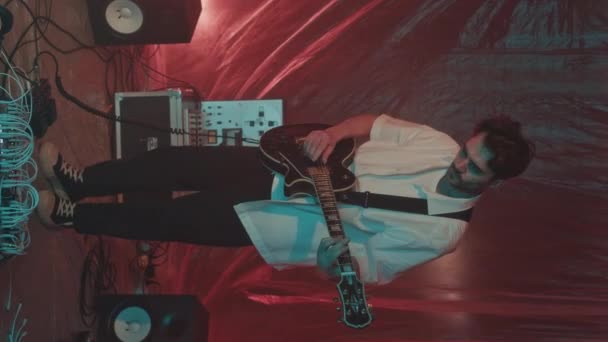 背の高い若い白人男性ギタリストの垂直フルスローモショット エレクトリックギターを調整しながら ポリエチレンシートの壁と床でネオンミュージックスタジオに立っています — ストック動画
