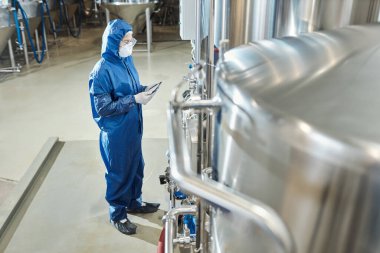 Endüstriyel kimya fabrikasında çelik tankların yanında koruyucu kıyafet giyen kadın işçiye yan bakış.