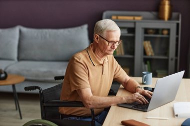 Tekerlekli sandalyede oturan ve masadaki dizüstü bilgisayarda yazan engelli bir adam.