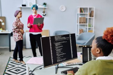 Siyahi genç bir kadının kod yazmasına ve ofiste genç yaratıcı bir ekiple bilgisayar kullanmasına yakın durun.