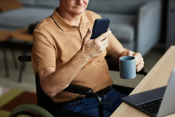 Close-up of senior man with disability sitting on wheelchair reading message on his smartphone and drinking coffee