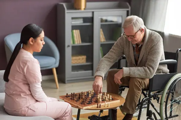 Senior man with disability playing chess with social worker while she visiting him at home
