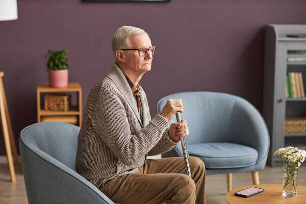Senior man with stick sitting on armchair with pensive sight, he spending time at home