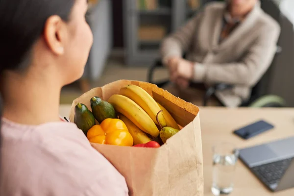 Rear view of social worker holding paper bag with fruits and vegetables, she buying food for senior man with disability