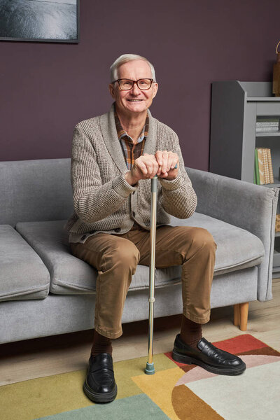 Portrait of senior man with stick smiling at camera while resting on sofa in living room