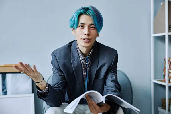 Minimal portrait of Asian young man with colored blue hair looking at camera holding documents in office and gesturing