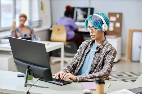 Portrait of young man with blue hair using computer in office and wearing headphones