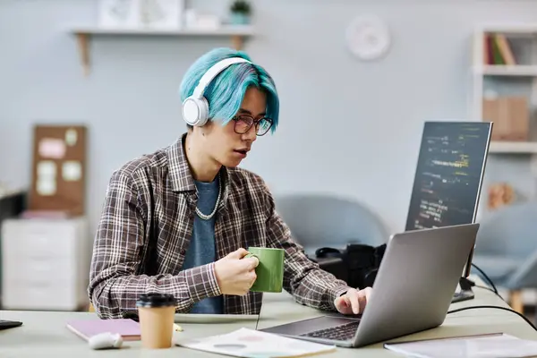 Portrait of young man with blue hair using computer in office and wearing headphones while writing code