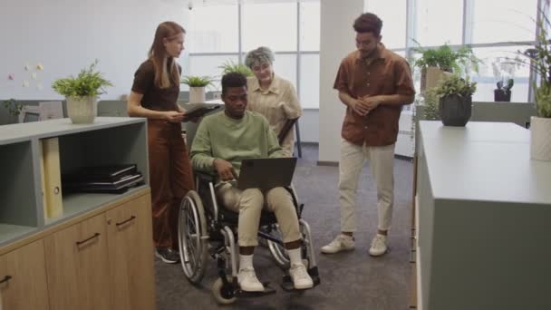 広々としたモダンなオフィスで クリエイティブな若い多様なチームの共同作業のフルショット 彼の太ももの上のラップトップと車椅子の黒人の男は同僚とプロジェクトのアイデアを共有する — ストック動画