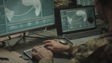 Kamuflaj üniformalı bir ordu subayının omzundan bilgisayar ve dizüstü bilgisayardaki uydu haritasına bakıyor, geceleri ofiste çalışırken mikrofonla kulaklık takıyor ve rapor gönderiyor.