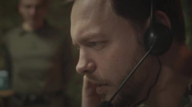 Kafkasyalı erkek askeri kontrol merkezi çalışanının, gece ofisindeki meslektaşlarıyla bilgisayardaki nesneleri takip ederken mikrofonla kulaklıkla rapor gönderdiği orta ölçekli bir görüntüsü.