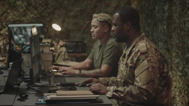 スクリーン上のCctvカメラのビューでコンピュータで座っている間 カメラのためにポーズする黒人軍将校の中程度のスローモの肖像画 Biracial女性の同僚との軍事コントロールセンターで働く — ストック動画