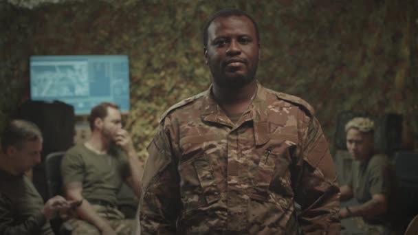 カムフラージュのユニフォームで若いアフリカ系アメリカ人男性の軍事監視官のミディアムスローモ肖像画は ぼやけた背景でブリーフィングをしている他の将校と折り畳まれた手で撮影します — ストック動画
