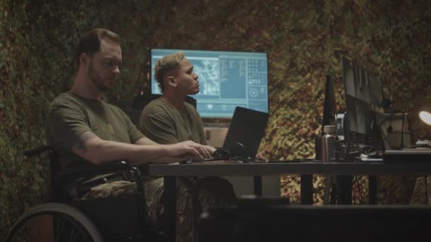 坐在轮椅上的年轻的白人军官和他的不同的男男女女同事在黑暗的办公室共同监督电脑屏幕上的监控程序 — 图库视频影像