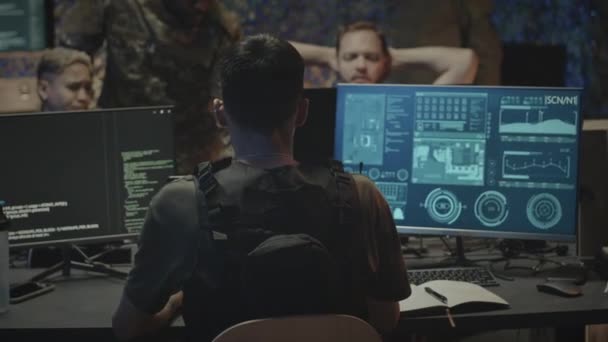 ダークオフィスのコンピュータ画面で監視プログラムの前に座っている男性のサイバーセキュリティ軍人労働者のバックビューショットを待って 監視とコントロールセンターの同僚と協力 — ストック動画