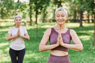 Açık havada yoga yapmaktan hoşlanan ve rahatlama egzersizi yaparken kameraya gülümseyen aktif son sınıf kadınının belden yukarı portresi.