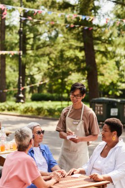 Gülümseyen genç garsonun dikey portresi açık kafede bir grup yaşlı kadına servis yapıyor.