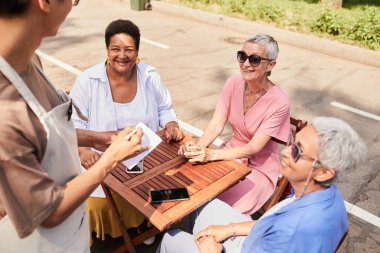 Gün ışığıyla aydınlatılan açık kafede sipariş veren bir grup neşeli son sınıf kadınına yüksek açılı bakış açısı