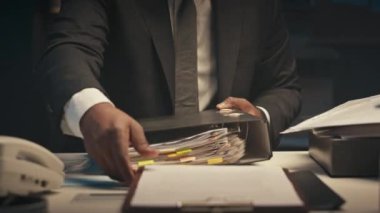 Siyah erkek girişimcinin gece yarısı karanlık bir ofiste gözlük, takım elbise ve kravat takıp, dosyalarla dolu kalın bir klasörü açarken ve incelemeye başlarken çekimini kaldır.
