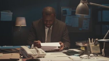 İşkolik Afrikalı Amerikan CEO 'nun beline zum yap evrak çalışırken kameraya bak, gece yarısı karanlık ofiste masa başında otur.