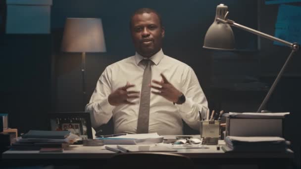 穿着白衬衫打着领带的有信心的黑人男性创业者 晚上坐在办公室的桌子旁 一边对着相机讲话 一边用手摆姿势 一边做产品演示 — 图库视频影像