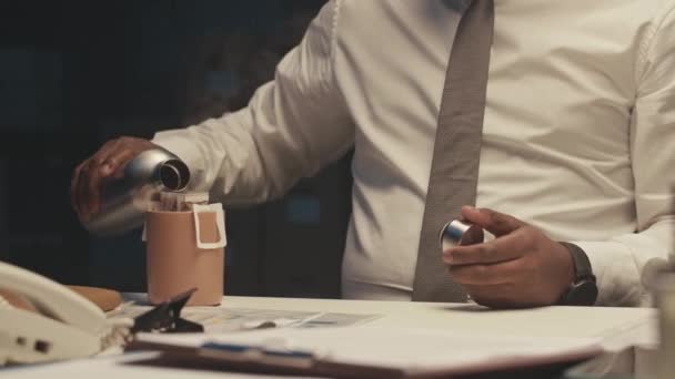 忙しいアフリカ系アメリカ人の男性起業家がメガネをかけ 暗いオフィスで机に座り 熱水を熱湯からカップに注ぎ 夜にコーヒーを飲んで働く — ストック動画