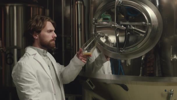 発酵タンクから新鮮なビールのサンプルを注ぎ その品質 質をチェックする白いラボコートのビールマスターの中型ショット — ストック動画