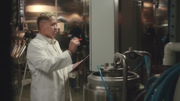 コントロールボードのボタンを押しながらクリップボードにメモを取るBiracial女性醸造技術者の中型ショット 醸造機器を検査 — ストック動画