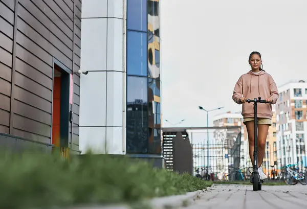 都市環境でのレジャーで現代の背の高い建物の列に沿って電動スクーターに乗るカジュアルな若い女子高生 — ストック写真