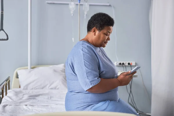 Minimal side view portrait of black senior woman using smartphone while sitting on bed in hospital room