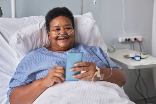 病院の部屋でベッドに横たわっている間 電話で笑顔のシニア女性の肖像 — ストック写真