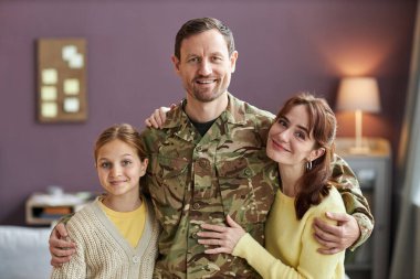 Waist up portrait of smiling military man embracing wife and daughter, all looking at camera in home setting clipart