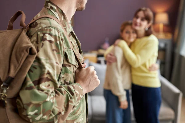 Close up of military man holding backpack looking at family in background, copy space