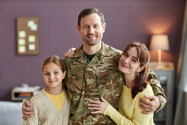 Waist up portrait of smiling military man embracing wife and daughter, all looking at camera in home setting