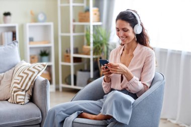 Gülümseyen genç bir kadının portresi. Hafta sonu evde eğleniyor ve kulaklıkla akıllı telefon kullanıyor.