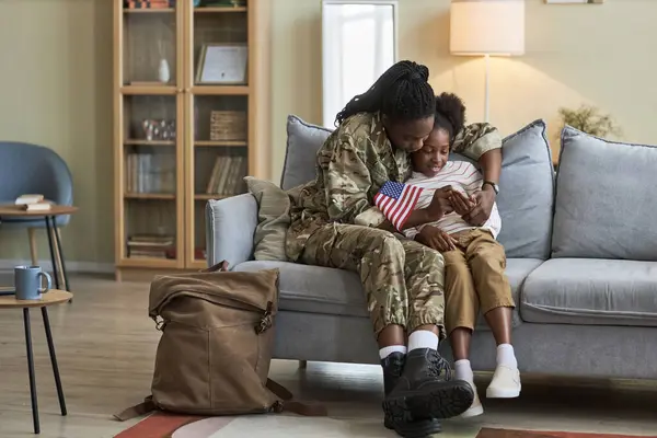 Young mother in military uniform returning home from war service, she sitting on sofa and talking with her little daughter
