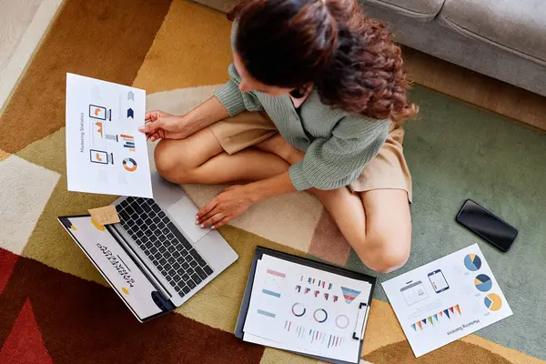 Top view portrait of woman working from home and using laptop while sitting on floor and analyzing data charts