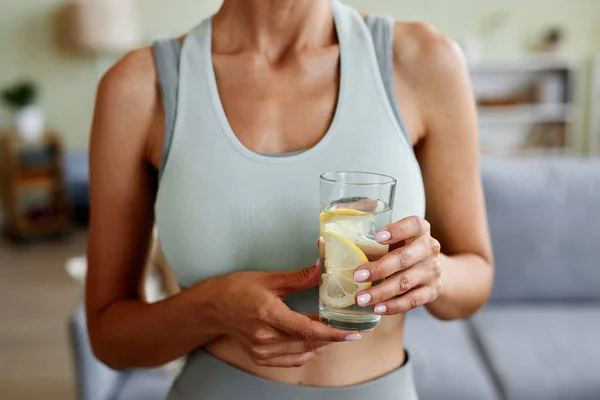 Cropped shot of sportive woman holding glass of water after home workout indoors