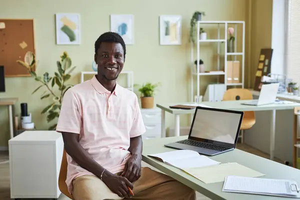 Portrait of young black man smiling at camera while sitting at workplace in cozy office, copy space