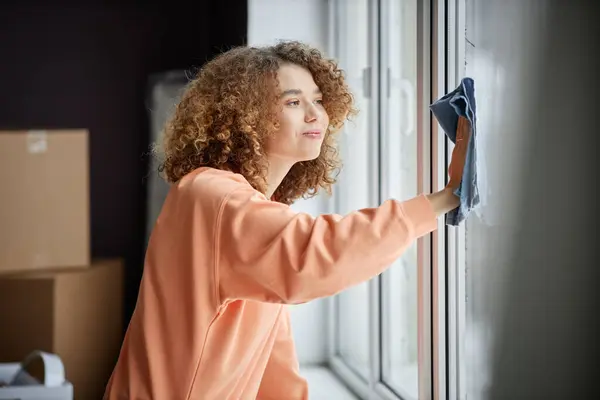 移転後に彼女の新しい家の窓から塵を拭くためにラグを使用する若い女性 — ストック写真