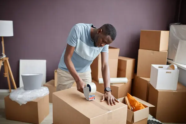 Young man packing boxes with adhesive tape during relocation in new home