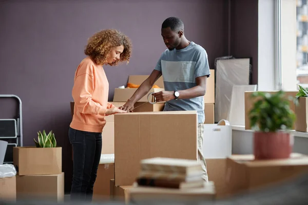 Young couple packing boxes together with adhesive tape during moving to a new house