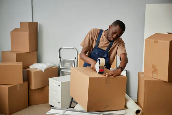 African American worker packing boxes with adhesive tape during relocation in new office