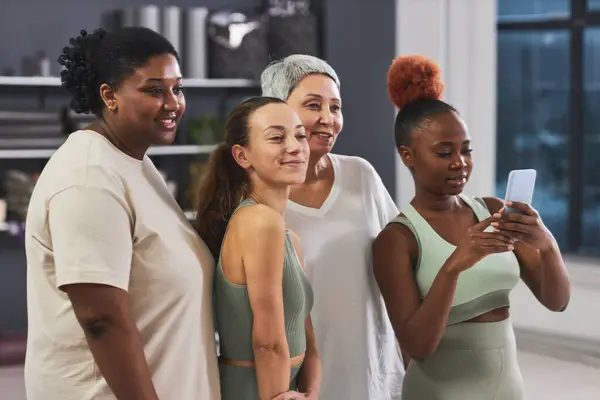 Group of multiethnic women posing on camera of smartphone together standing in gym