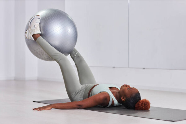 Афроамериканская молодая женщина, лежащая на коврике для упражнений и тренирующаяся с мячом