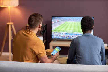 Televizyonda futbol maçı izleyen ve elinde online bahislerle akıllı telefon tutan iki adamın arka planı.