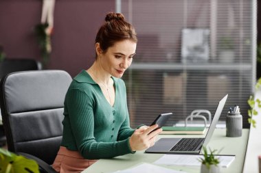 Ofiste çalışırken masasında yeşil bitkilerle süslenmiş akıllı telefon kullanan gülümseyen genç bir kadının yan görünümü