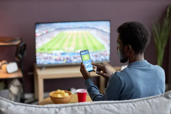 男子手持智能手机的背景图 屏幕上有体育投注应用程序 同时在家中观看足球比赛 复制空间 — 图库照片
