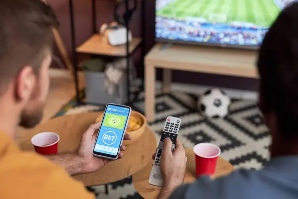 两名男子在电视上观看足球比赛 并拿着智能手机与网上体育赌博 — 图库照片
